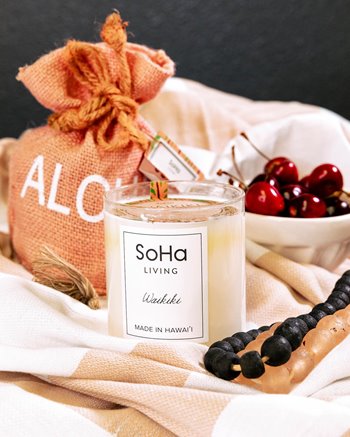 Soha Living candle next to a bowl of cherries & an orange bag that says aloha on it.