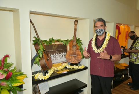 Man wearing a flower lei standing next a Hawaiian music display with 2 guitars & a black-and-white photo.