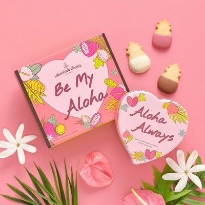 Pink "Be My Aloha" Box & "Aloha Always" heart-shaped tin from Honolulu Cookie Company set against a pink backdrop. There are pink tropical flowers & green leaves at the bottom of the shot as well as 3 pineapple-shaped cookies in the upper-right corner.