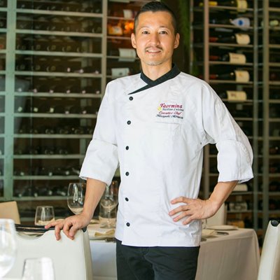 Hiroyuki Mimura, Executive Chef at Taormina Sicilian Cuisine, wearing a white executive chef's coat & posing in front of a dining table & clear wine cabinet.