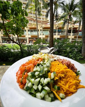 Cobb salad with diced cucumbers, tomatoes, egg, cheese, & meat in a plate with a view of Waikiki Beach Walk shops in the background.
