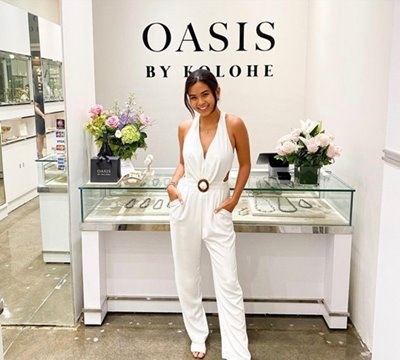 Rose Wong, CEO and Founder of Kolohe, wearing a white jumpsuit & posing with hands in her pockets in front of a wall saying "Oasis by Kolohe" with a jewelry display case underneath.