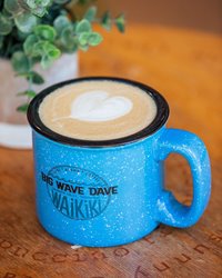 Blue cup with the words "Big Wave Dave Waikiki" on it & latte art in the form of a heart on a wooden table with a plant in the back.