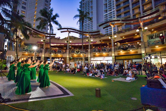 Group of hula dancers wearing long green dresses performing for the crowd in front of Waikiki Beach Walk