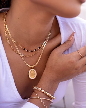 Woman in white wearing 3 layered gold necklaces. Two of the necklaces are chains that sit closer to the neck and collarbone, while the third necklace features a thicker herringbone-style chain with a round pendant. She also wears a gold crisscrossed bracelet and gold beaded bracelet on one hand.