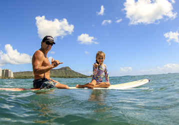 Adult man & young girl putting up shakas on a surfboard in the water with Diamond Head in the background.