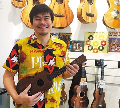 Tyler Gilman, Owner of The Ukulele Store, in a yellow collared shirt & posing with an ukulele in hands while standing in front of a wall with ukulele & guitars hanging.