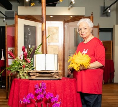 Antoinette Toni Lee in a red shirt, standing next to an open book propped up on a table while holding yellow feathers.