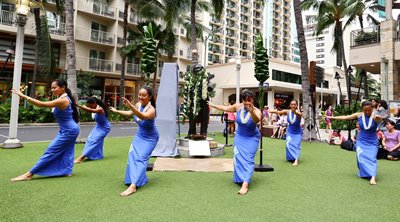 Hula troupe wearing blue dresses, in a dance pose around the Gabby Pahinui statue