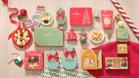 A collection of different red & green holiday-themed cookie boxes. Pineapple-shaped cookies of various flavors are plated in different areas of the photo.