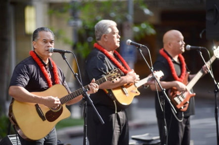 3 men in black shirts, each playing a guitar & singing in front of a microphone while performing in Waikiki
