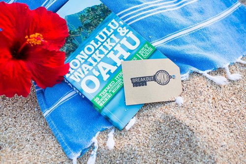 Breakout Waikiki card on top of a guidebook that says Honolulu, Waikiki, & Oahu, which is laying on a blue towel on the sand next to a red hibiscus flower.