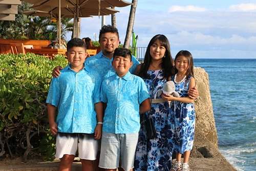 Father & two sons in matching aqua blue aloha shirts standing next to the mother & daughter wearing blue floral sundresses - all smiling for a photo by the ocean