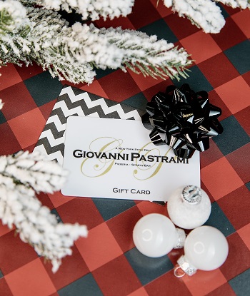 Giovanni Pastrami Gift Card with a black bow on top, with a red and black checkered cloth underneath & snow-frosted tree branches around it.
