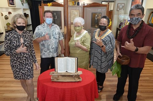 Group of people standing around an open book on display with various other Hawaiian cultural artifacts in The Royal Room at Waikiki Beach Walk.
