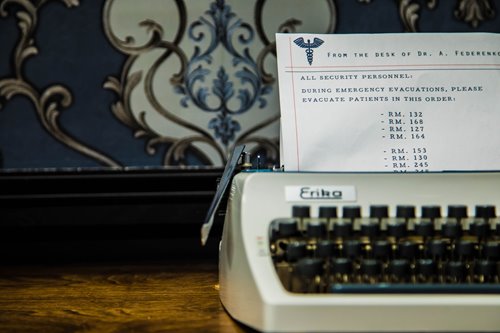 White typewriter with a paper reading "FROM THE DESK OF DR. A. FEDERENK. ALL SECURITY PERSONNEL: DURING EMERGENCY EVACUATIONS, PLEASE EVACUATE PATIENTS IN THIS ORDER:" followed by a list of room numbers
