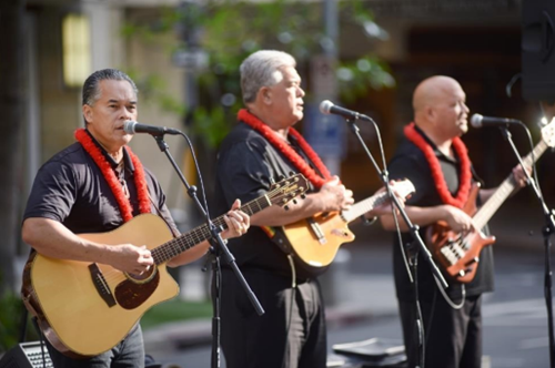 3 local musicians, each wearing red lei & holding guitars while singing into microphones