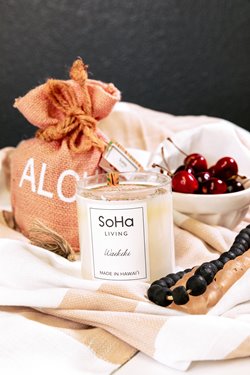 Soha Living candle next to a bowl of cherries & an orange bag that says aloha on it.