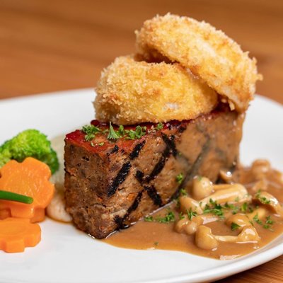 Square meatloaf with 2 fried rings on top, side of vegetables, & mushroom sauce on a plate.