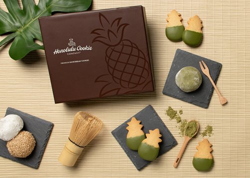 Brown Honolulu Cookie Company box next to cookies dipped in green matcha-infused chocolate, green tea powder, & mochi.