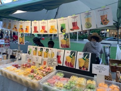 Pictures of different smoothie flavors hanging at a farmers market booth with fresh fruit packaged underneath