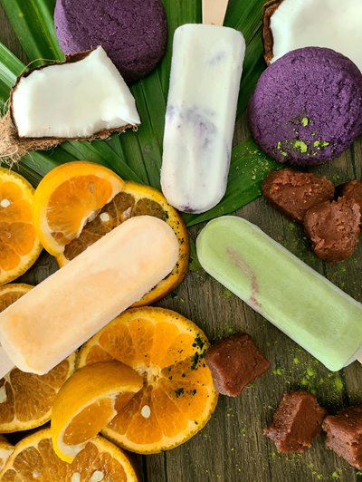 White & purple, pastel orange, and pastel green popsicles next to an open piece of coconut, ube cookie, & orange slices.