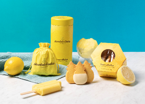 Honolulu Cookie Company's lemon flavor collection consisting of a yellow window box, cylindrical tin, & drawstring bag next to 3 pineapple-shaped cookies, lemons, and a yellow popsicle against a teal blue background.
