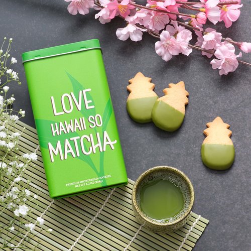 Green tin box saying "Love Hawaii So Matcha" next to green, matcha-dipped pineapple-shaped shortbread cookies. Around these two items are cherry blossom petals, a cup of green tea, & white flowers.