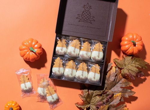 Open box of Honolulu Cookie Company's white chocolate pumpkin treats next to mini pumpkins, fallen leaves & 3 cookies laid out on an orange surface next to the box.