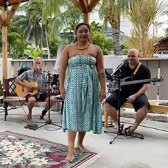 Woman in a teal and while dress preparing to dance hula while 2 musicians play behind her.