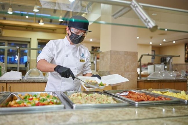 An employee wearing a face mask, face shield, & gloves packing food into a to-go container at the breakfast buffet.