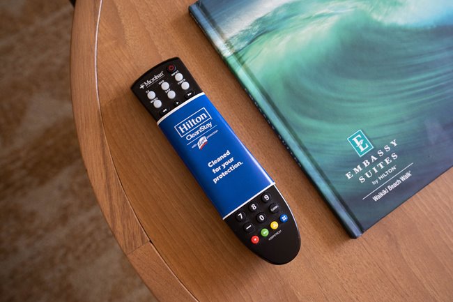 A remote control wrapped in a Hilton CleanStay label saying "Cleaned for your protection" placed on a wooden table next to an Embassy Suites Waikiki book.
