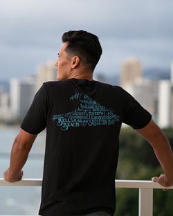 Man on a balcony looking out towards the ocean while wearing a black tea with a wave-shaped graphic made of words on the back.