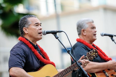 Two men wearing black shirts & red lei, each playing the guitar while singing into a microphone.