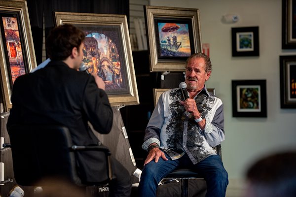 Artist James Coleman holding a microphone while speaking to a man in a black collared shirt. Coleman's paintings can be seen next to them in frames.