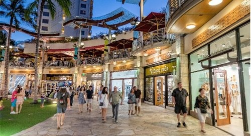 A crowd of people walk in front of the first floor stores at Waikiki Beach Walk