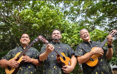3 men smiling while wearing matching black aloha shirts with floral outlines & holding ukuleles while standing under the trees.