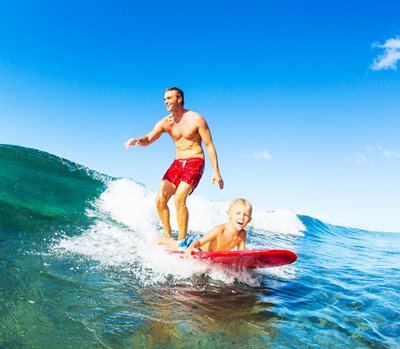 Man surfing a wave with his son laying at the front end of the board