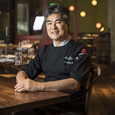 Professional photo of Roy Yamaguchi in a black chef's coat, sitting at a table & smiling at the camera.