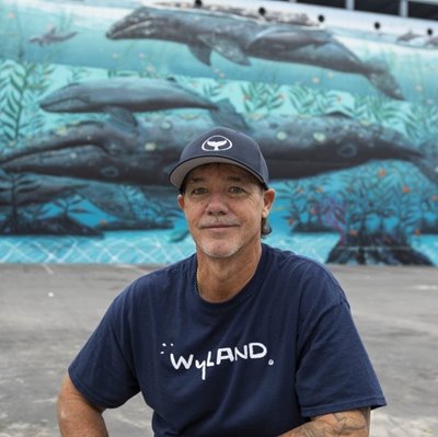 Wyland is wearing a navy blue hat donning a whale's tail fin logo & matching navy blue shirt with the word "Wyland" on it, while squatting in front of a marine life mural depicting whales in the ocean.