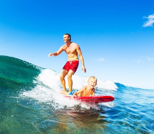 Man standing up on the back of a surfboard with a young boy lying on the front while riding a wave.
