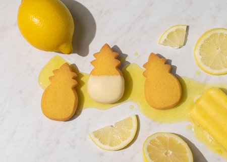 3 lemon-flavored cookies in the shape of a pineapple laying on marble next to slices of lemon and a yellow popsicle.