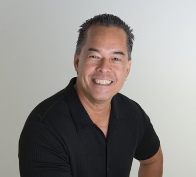 Photo of Blaine Kia, Entertainment Director and Cultural Advisor at Waikiki Beach Walk, in a black collared shirt & smiling directly at the camera