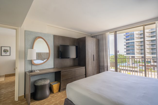 A look at the inside of a room at Embassy Suites Waikiki Beach Walk showing a King bed, mirror, TV, & balcony with partial ocean view
