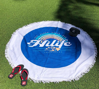 White & blue round towel with HiLife and a rainbow at the center on a patch of grass.