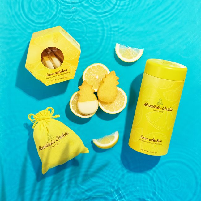 2 pineapple-shaped lemon cookies sitting on 3 lemon slices surrounded by Honolulu Cookie Company's yellow box, tin, and bag packages
