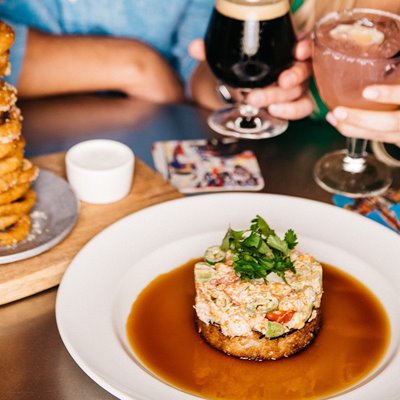 The Cali Stack appetizer on a white plate with a dark beer & pink cocktail being held in the background.