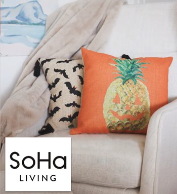 Two pillows on couch. The pillow in front has a design that features an orange background and a pineapple that is carved like a Halloween pumpkin. The pillow behind it has a bat-pattern design with black tassels on the corners. The picture is overlaid with a logo that reads "SoHa Living."